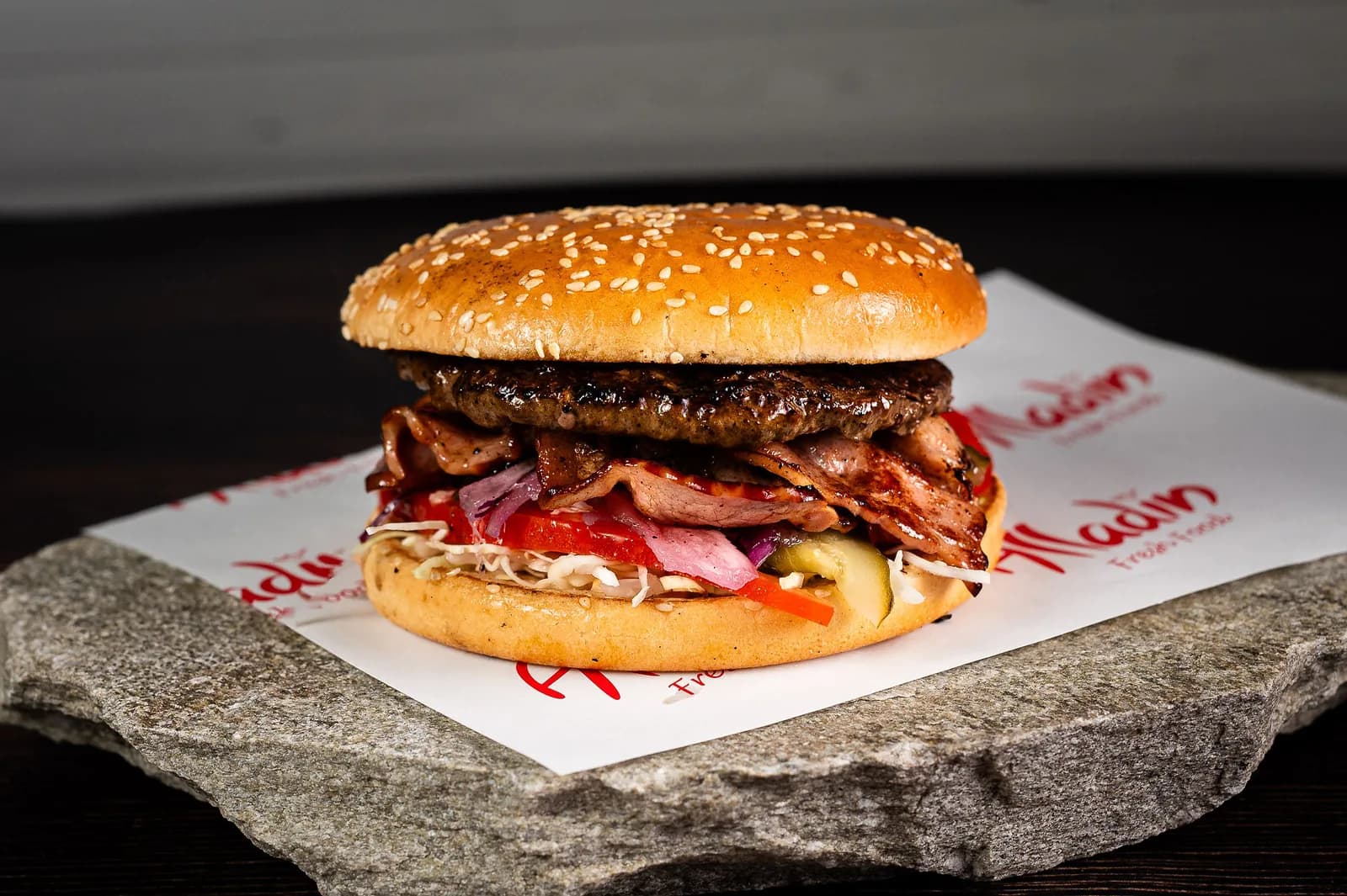 Bacon Burger: chiflă cu susan, carne de vită, bacon, salată de varză, roșii, castraveți murați și ceapă roșie, pe hârtia Alladin