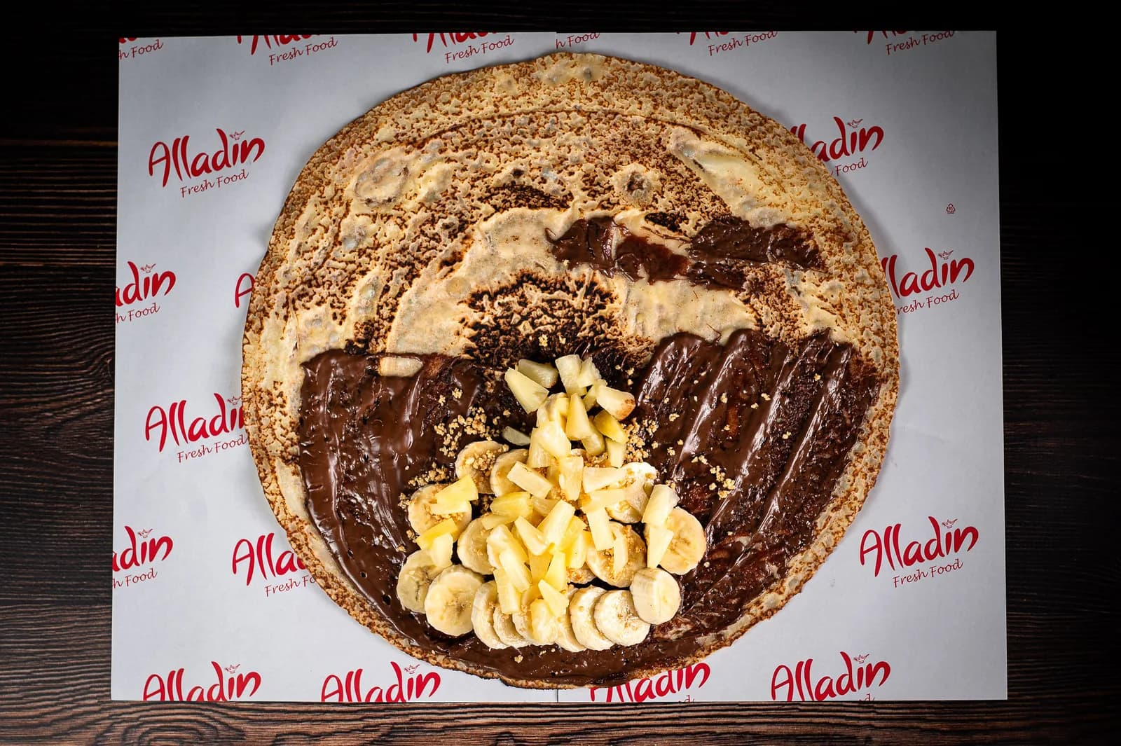 Clătită Fresh desfăcută: Nutella, banane, ananas și arahide măcinate, pe hârtia brandată Alladin